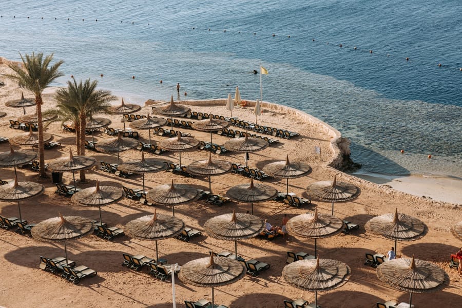 Vista aerea della spiaggia sabbiosa di Reef Oasis con file di lettini e ombrelloni di paglia vuoti, delimitata da palme e dal mare calmo di Blue Bay. Alcune persone nuotano e si rilassano vicino alla riva.