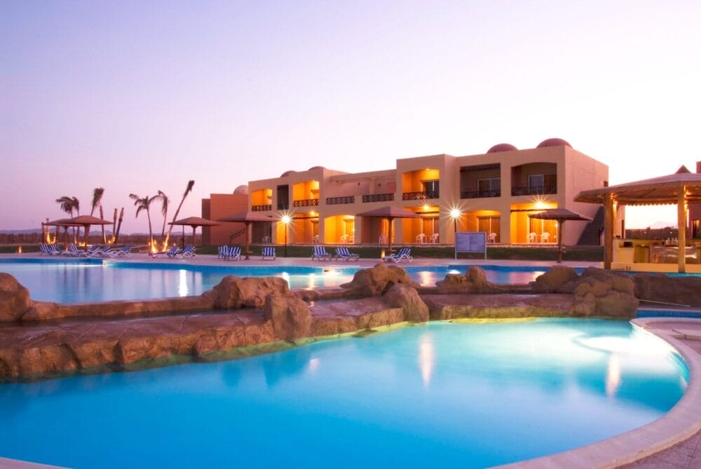 Un moderno edificio del resort con balconi si affaccia su una grande piscina all'aperto al tramonto; palme, sedie a sdraio e cabine a bordo piscina del Wady Lahmy Azur Resort creano un'atmosfera di vacanza rilassata.