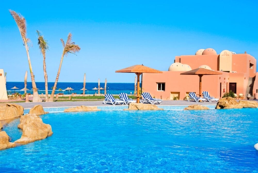 Una piscina blu brillante con sedie a sdraio e ombrelloni, palme e un edificio color terracotta al Wady Lahmy Azur Resort vicino all'oceano in una giornata di sole.