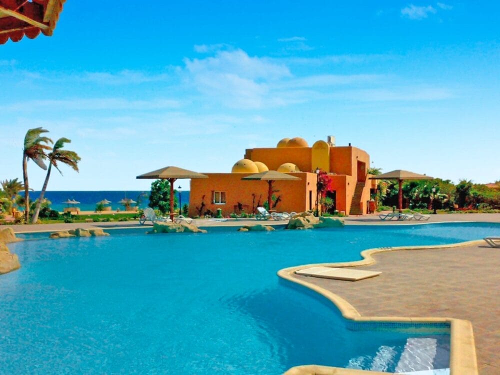 La grande piscina all'aperto con bordi curvi del Wady Lahmy Azur Resort si affaccia sul mare; le palme ondeggiano nella brezza accanto a un edificio arancione a cupola, sotto un cielo blu brillante. Sedie a sdraio e ombrelloni circondano la piscina.