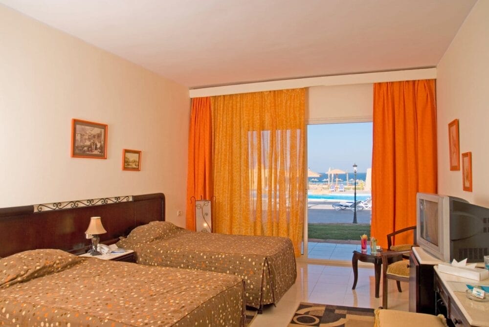 Una camera d'albergo del Wady Lahmy Azur Resort con due letti singoli, tende arancioni, opere d'arte incorniciate sulle pareti color crema, una TV su una scrivania e una vista sulla piscina e sull'area salotto all'aperto attraverso una grande porta a vetri.