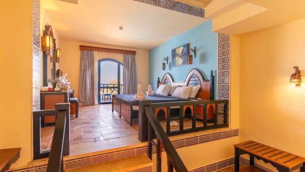 Accogliente camera dell'hotel Faraana Heights con due letti, piastrelle colorate, una parete blu e un balcone con tende. Un'illuminazione calda, una scrivania, una panca e opere d'arte decorative contribuiscono a creare un'atmosfera invitante.