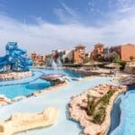 Una grande piscina di un resort con scivoli, rocce e fontane sotto il cielo azzurro di Faraana Heights. Nell'angolo in alto a sinistra, il logo "Italian Escape" con un disegno verde, bianco e rosso.