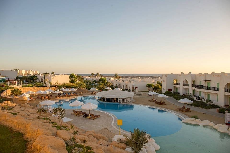 L'Hilton Nubian Village dispone di una grande piscina curva circondata da sedie a sdraio e ombrelloni bianchi, con edifici bianchi e palme sullo sfondo, sotto un cielo limpido al tramonto.