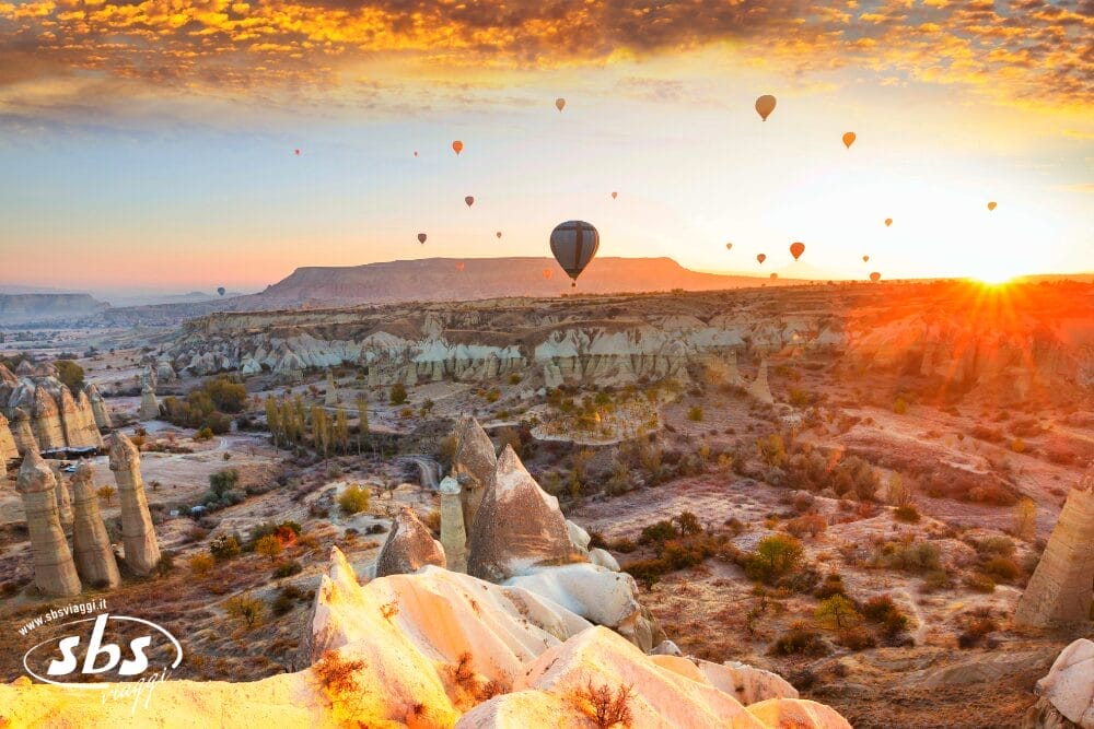 Mongolfiere fluttuano sopra una valle rocciosa e panoramica all'alba in Cappadocia, Turchia. Scoprite questa vista mozzafiato durante un mini tour, mentre formazioni rocciose uniche e un cielo spettacolare sono immersi nella calda luce dorata del sole.