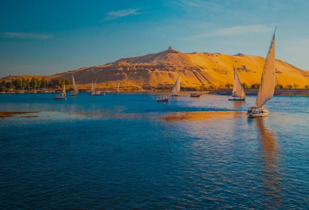 Barche a vela con vele bianche scivolano su un fiume blu vicino a Luxor, con colline di sabbia dorata sullo sfondo sotto un cielo limpido: uno scenario perfetto per un'avventura di Crociera Ramses.