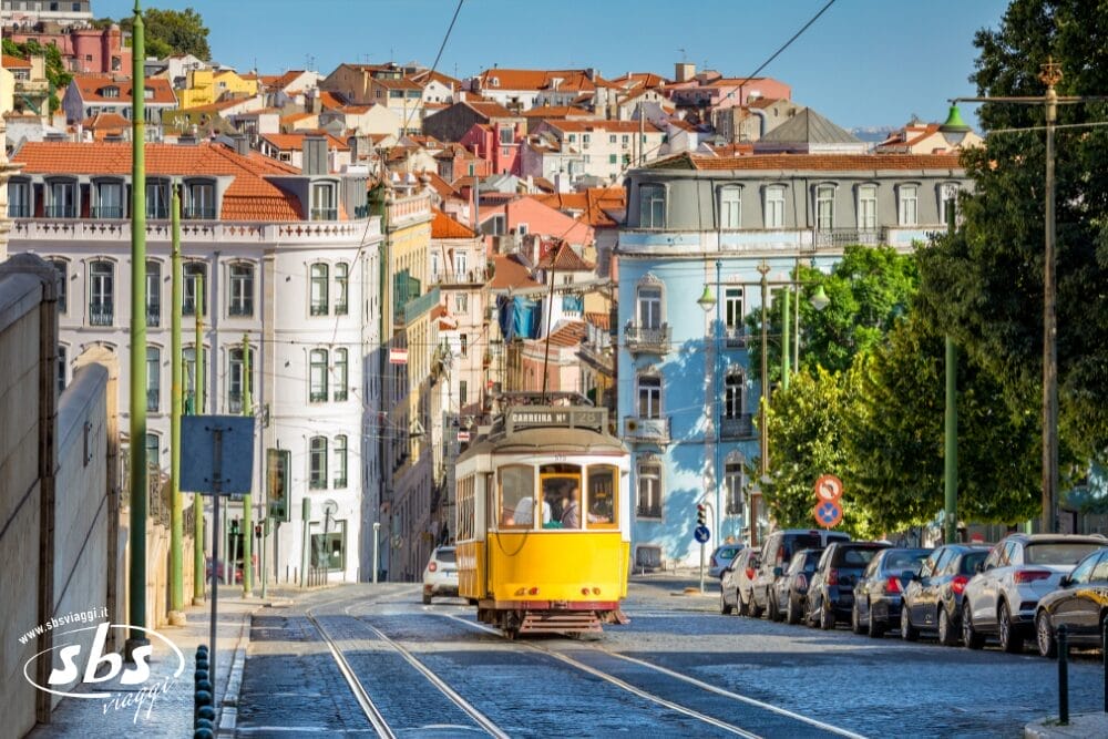 Un tram giallo viaggia lungo i binari di una strada soleggiata di Lisbona, Portogallo, passando davanti a edifici colorati e auto parcheggiate. Il paesaggio collinare della città e il cielo azzurro fanno da cornice a un affascinante minitour durante la festa locale.