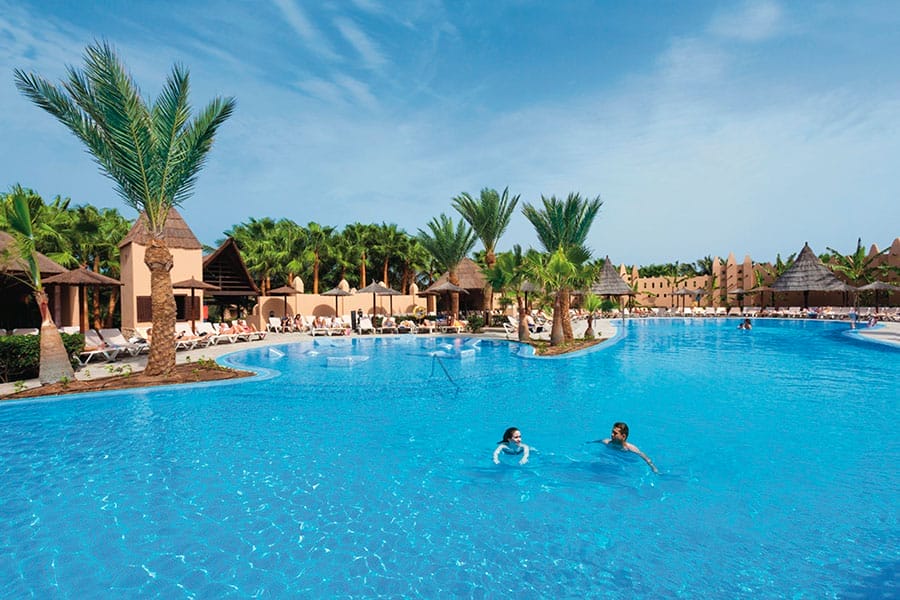 Grande piscina all'aperto con due persone in acqua, circondata da palme, sedie a sdraio e tetti di paglia sotto un cielo azzurro e limpido.