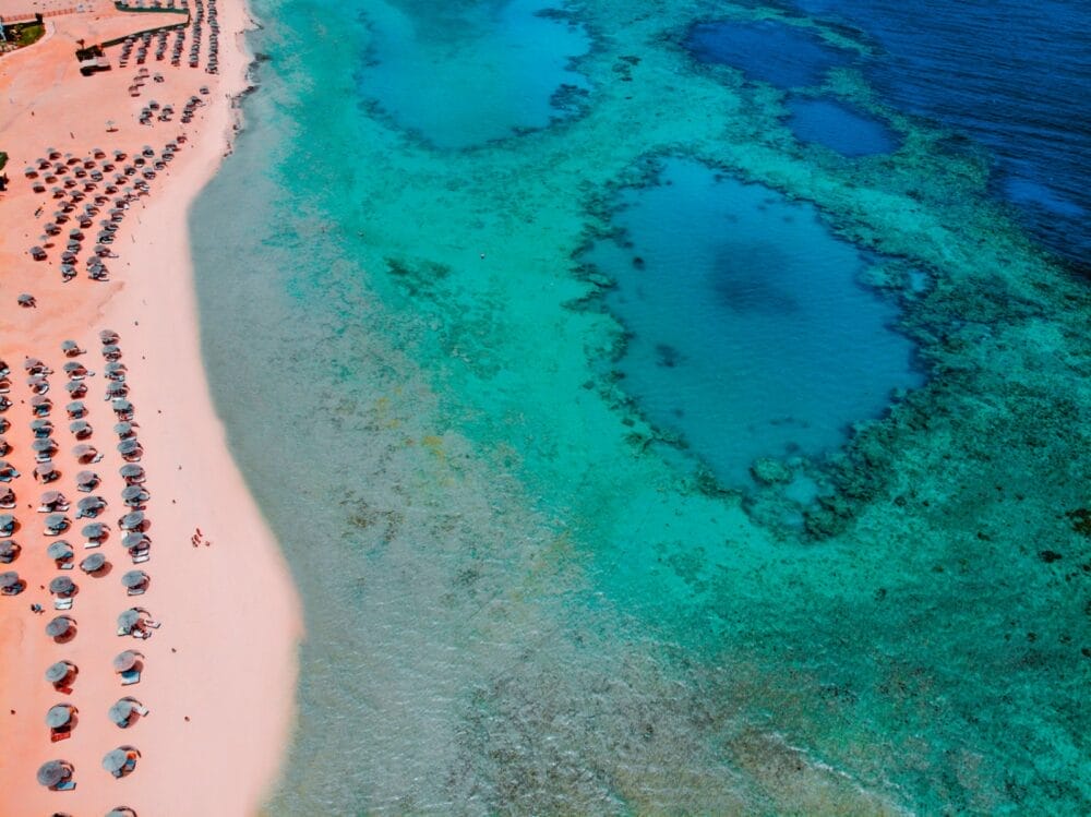 Veduta aerea della spiaggia di Gorgonia con file di lettini e ombrelloni sulla sabbia rosata, accanto a un'acqua limpida e turchese e alla barriera corallina visibile, perfetta per un soggiorno rilassante al Seaclub Gorgonia Beach Resort.