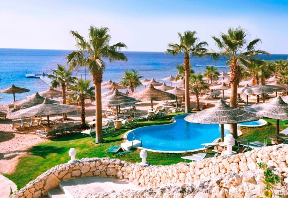 Un resort tropicale sulla spiaggia di Sharm El Sheikh con palme, sedie a sdraio, ombrelloni di paglia, una piscina e una vista sul mare blu e calmo in una giornata di sole.