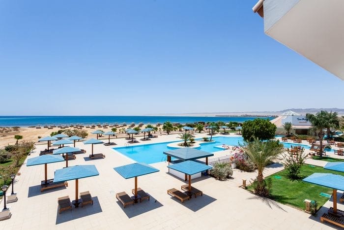 Il Seaclub Lahami Bay è un resort con una grande piscina all'aperto, lettini sotto pergolati blu, palme e prati verdi, che si affaccia su una spiaggia sabbiosa e sul mare blu sotto un cielo limpido.