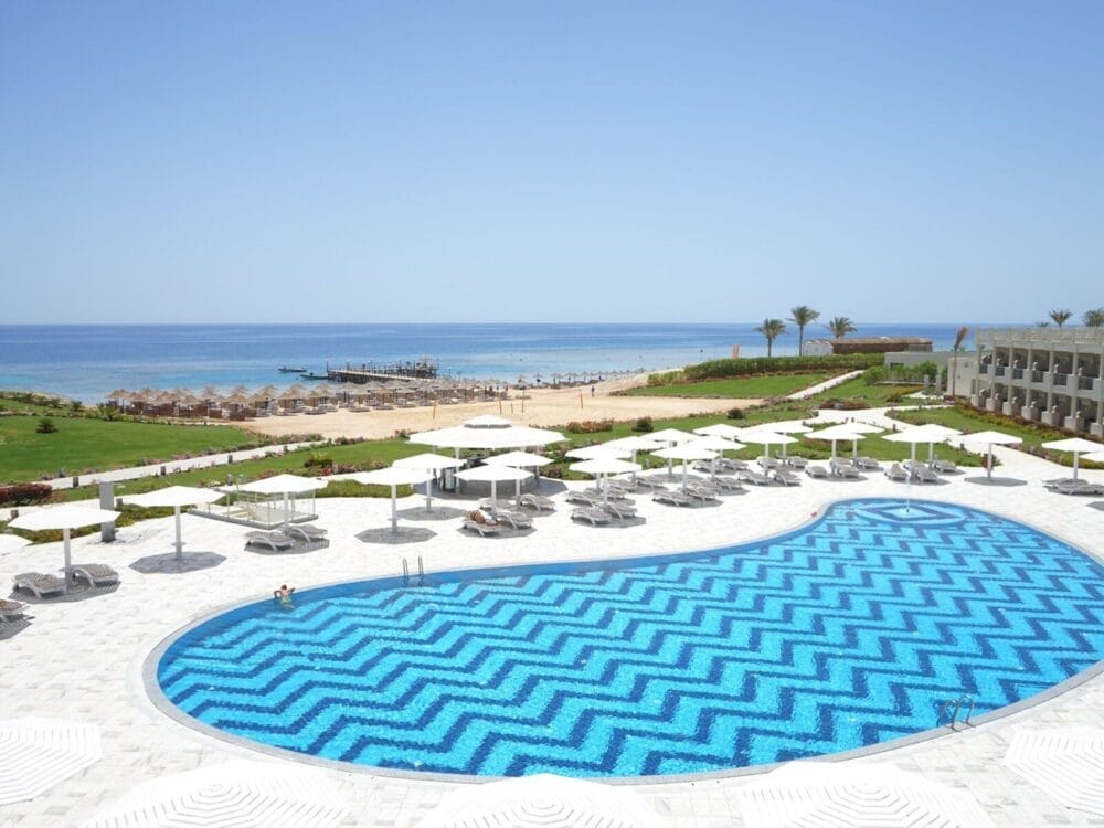 Il Seaclub Sirena Beach Resort dispone di un'ampia piscina con un motivo a zig zag blu, circondata da sedie a sdraio e ombrelloni bianchi, che si affaccia sulla spiaggia sabbiosa e sul mare calmo sotto un cielo azzurro e limpido.