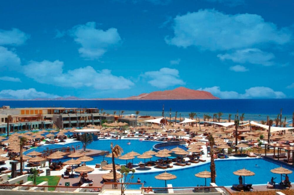 Il Coral Sea Imperial Searesort dispone di diverse piscine blu, lettini e ombrelloni di paglia con vista sul mare. Sullo sfondo, un'isola riposa sotto un cielo blu brillante punteggiato da nuvole sparse.