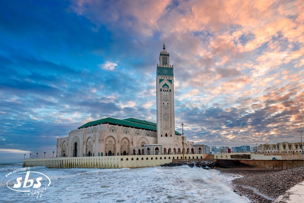 La Moschea di Hassan II si erge in riva all'Oceano Atlantico a Casablanca, in Marocco, con il suo alto minareto e la sua intricata architettura ispirata ai Borghi Andalusi, sotto un cielo vibrante e pieno di nuvole al tramonto, mentre le onde si infrangono sulla vicina riva rocciosa.