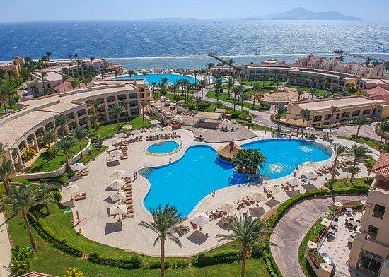 Veduta aerea del Cleopatra Luxury Resort con grandi piscine sinuose, lettini, palme ed edifici circostanti, affacciati sul mare con montagne lontane sotto un cielo limpido.