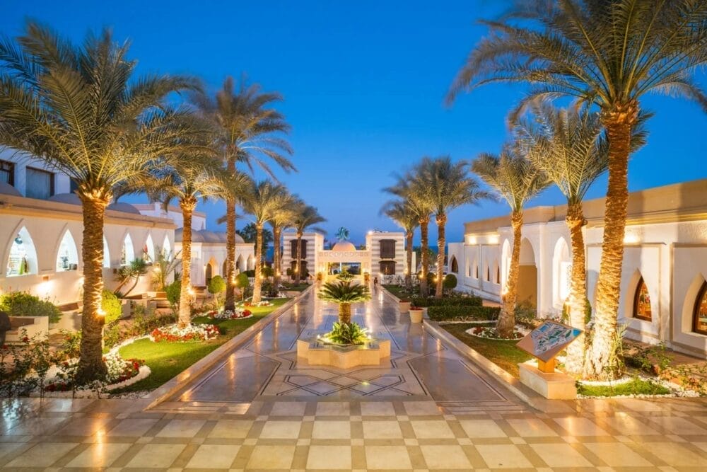 Un lussuoso cortile del Reef Beach Resort con palme, camminamenti illuminati, aiuole ed edifici bianchi ad arco su entrambi i lati, sotto un cielo sereno, perfetto per i raduni del Bravo Smart Club.