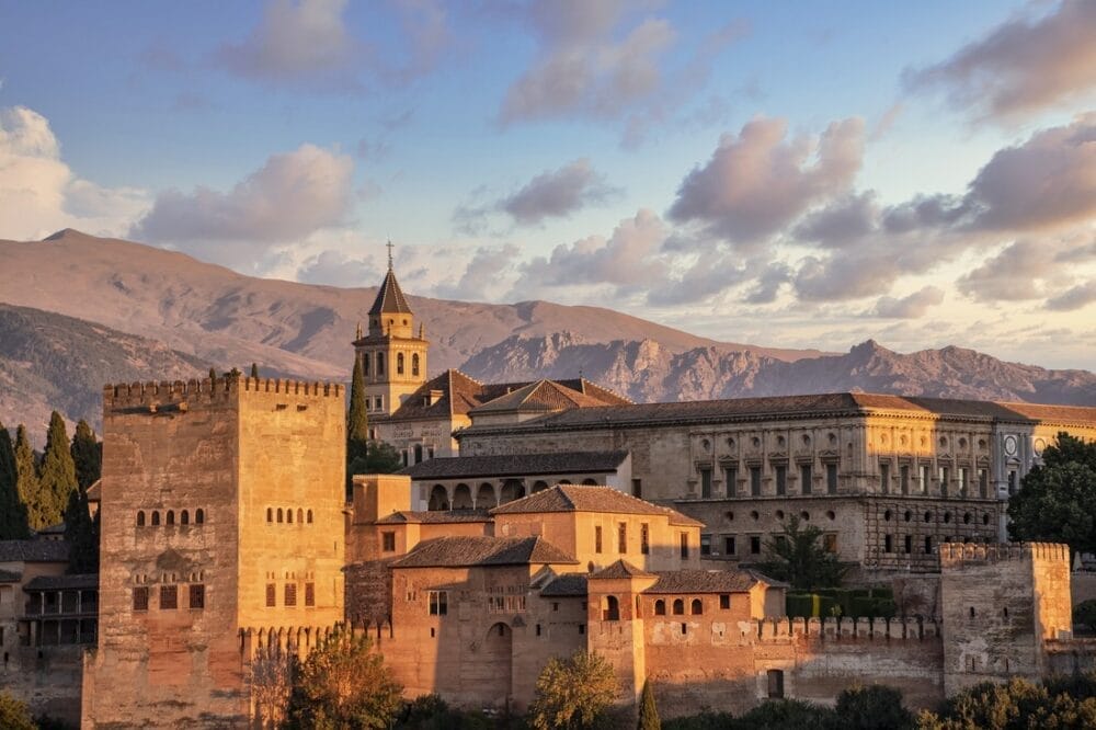 Il complesso del palazzo dell'Alhambra a Granada, in Spagna, viene visto al tramonto durante il Gran Tour Andalusia, con le sue torri e le sue mura storiche immerse in una luce calda, sullo sfondo delle montagne e di un cielo parzialmente nuvoloso: un'esperienza indimenticabile per Capodanno 2025.