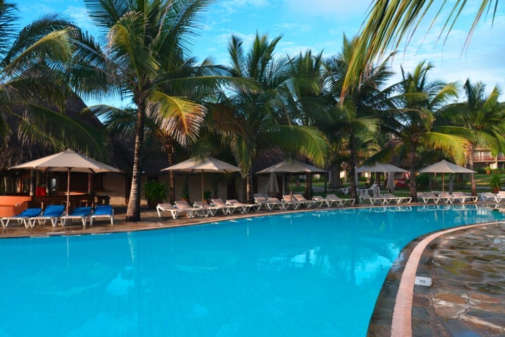 Una piscina del resort circondata da palme, lettini e ombrelloni beige. L'acqua cristallina e l'atmosfera pacifica e tropicale creano la fuga perfetta del Seaclub sotto un cielo parzialmente nuvoloso.