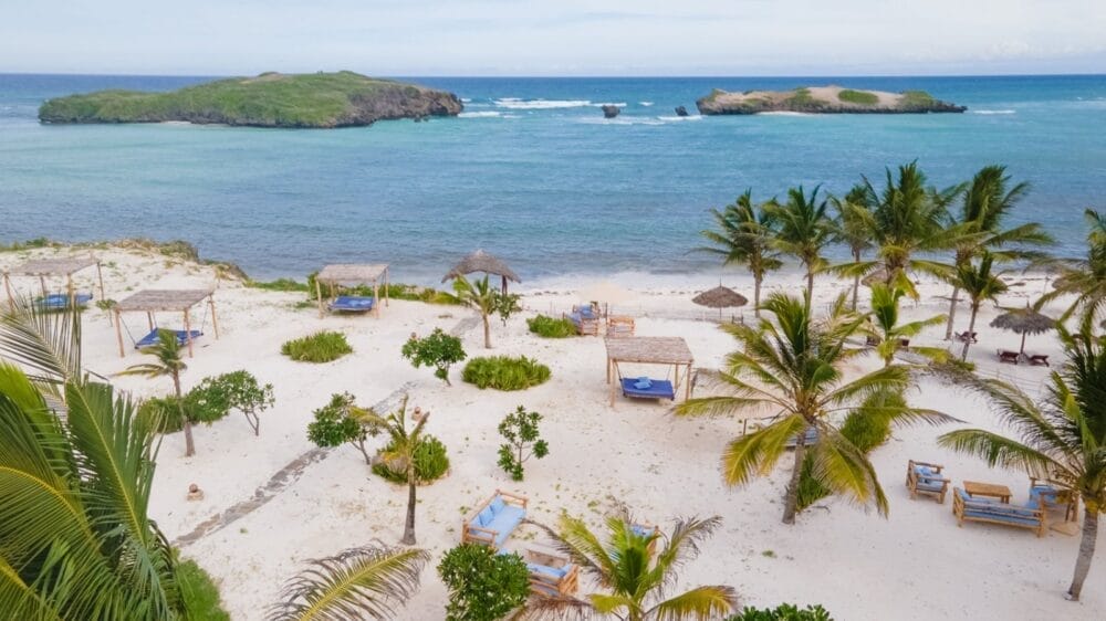 Vista aerea di una spiaggia tropicale con sabbia bianca, acqua cristallina, palme e cabine in legno. Piccole isole verdi sono visibili in lontananza sotto un cielo parzialmente nuvoloso al Seaclub 7 Island Resort.