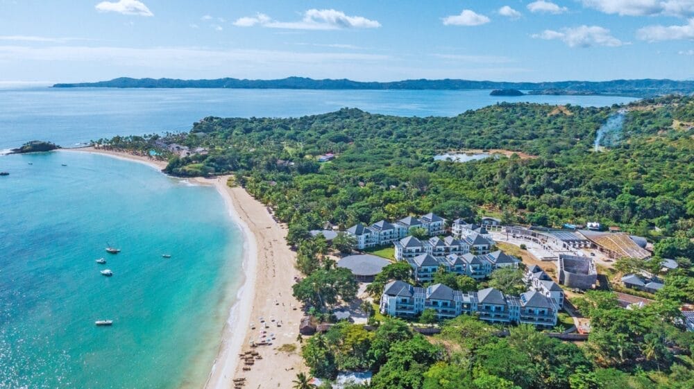 Vista aerea del Seaclub Royal Andilana, resort sulla spiaggia con acqua turchese, sabbia bianca e alberi verdi. Rilassatevi nella spa mentre le colline e la costa si estendono in lontananza sotto un cielo blu con nuvole sparse.