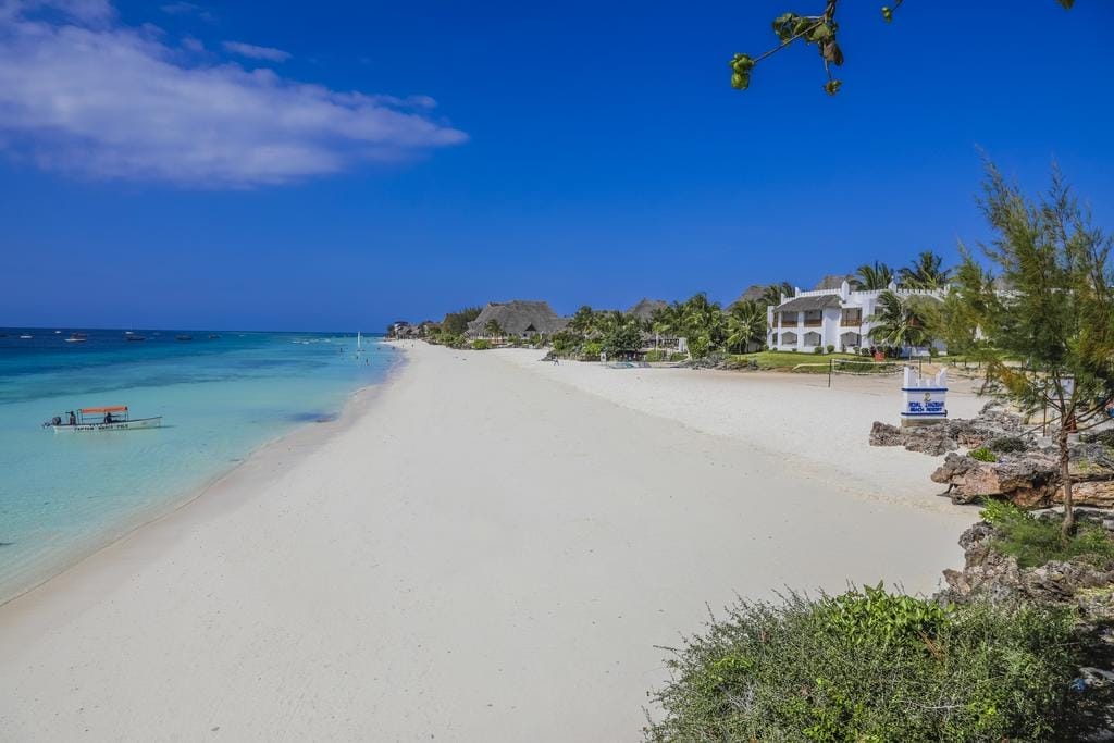 Un'ampia spiaggia sabbiosa di Zanzibar con acqua turchese e limpida, piccole imbarcazioni al largo, alberi verdeggianti ed edifici bianchi in riva al mare sotto un cielo blu brillante.