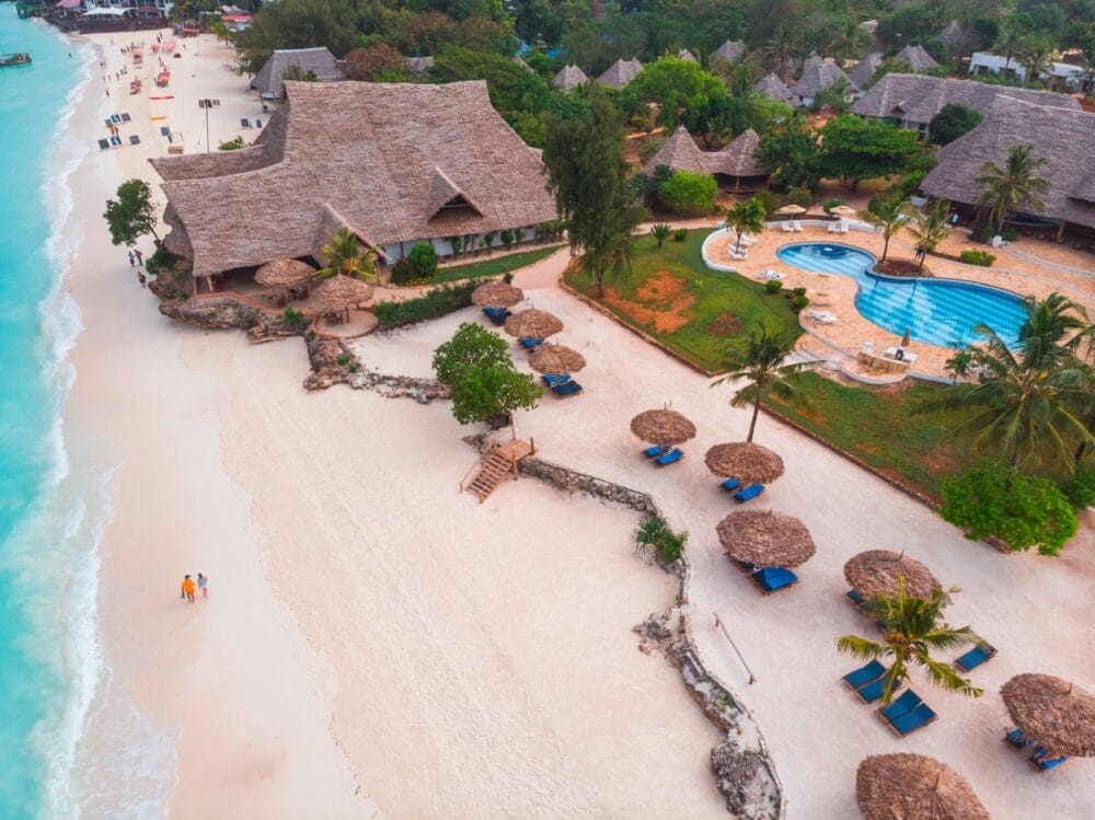 Vista aerea del Seaclub Sandies Baobab a Zanzibar, con edifici dai tetti di paglia, una piscina sinuosa, sedie a sdraio sotto ombrelloni di paglia e onde turchesi dell'oceano su sabbia bianca. Due persone camminano lungo il litorale.