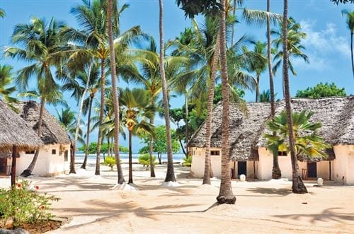 Capanne dal tetto di paglia circondate da alte palme sulla spiaggia sabbiosa del Seaclub Diamond Mapenzi Beach, con il cielo azzurro e l'oceano sullo sfondo.