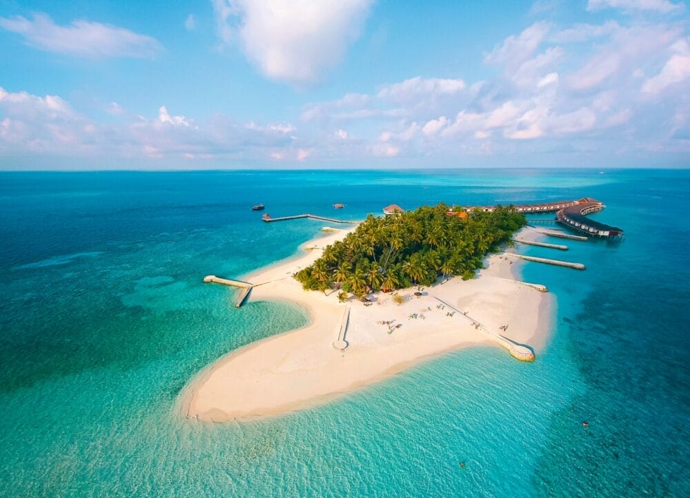 Vista aerea di una piccola isola tropicale, sede del Seaclub Dhiggiri, con spiagge di sabbia bianca, palme verdi e acque blu turchese, circondata da pontili e ville overwater: l'ideale per una vacanza al mare in un resort delle Maldive sotto un cielo parzialmente nuvoloso.