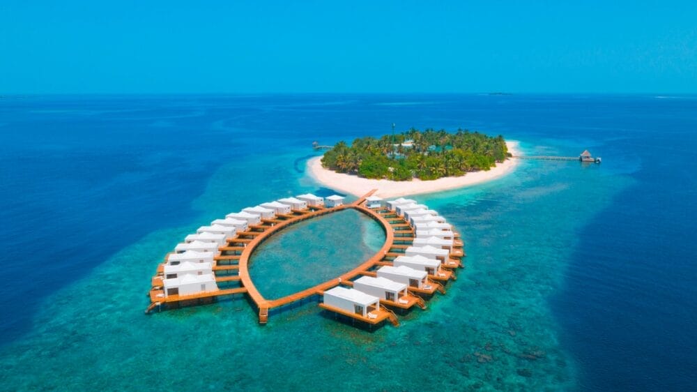Vista aerea del resort Sandies Bathala, un'isola tropicale lussureggiante circondata da acque turchesi, con bungalow overwater disposti a semicerchio e collegati da passerelle di legno che riportano al Seaclub.