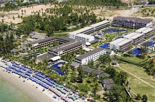 Vista aerea del Sunprime Kamala Beach Resort, un resort marittimo con edifici bianchi, palme lussureggianti, piscine e file di sedie a sdraio blu che costeggiano la spiaggia sabbiosa di Kamala, accanto all'acqua cristallina.