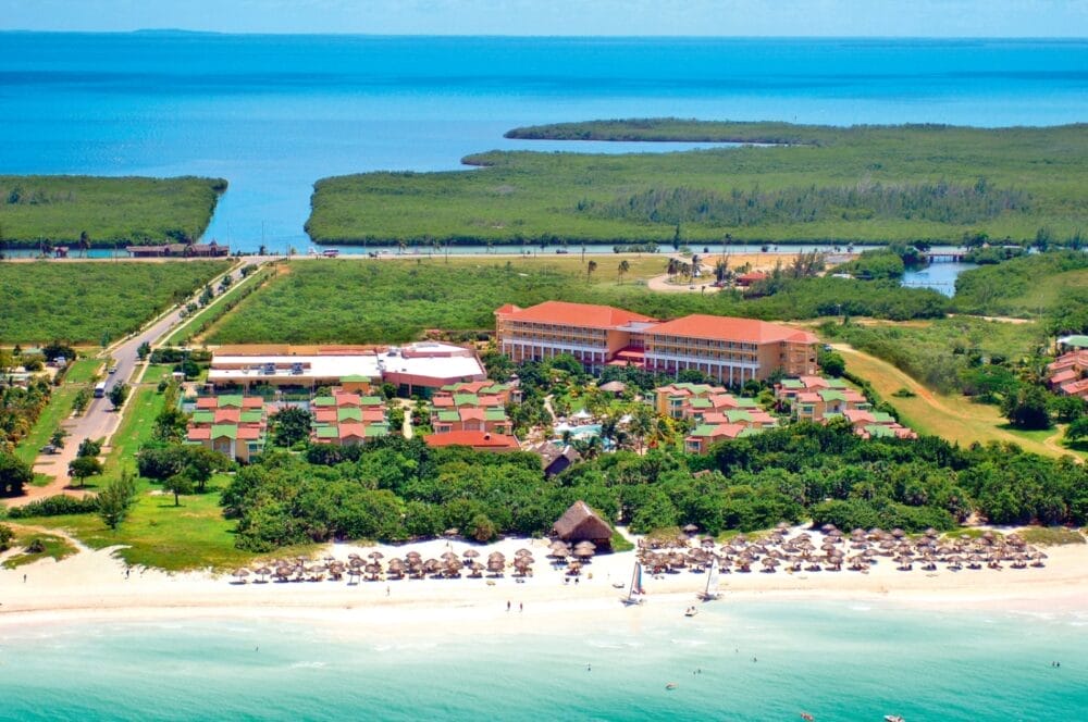 Vista aerea del resort Iberostar Tainos Seaclub con edifici dai tetti rossi, palme, litorale sabbioso, ombrelloni di paglia e oceano turchese; sullo sfondo un paesaggio verde e acque calme.