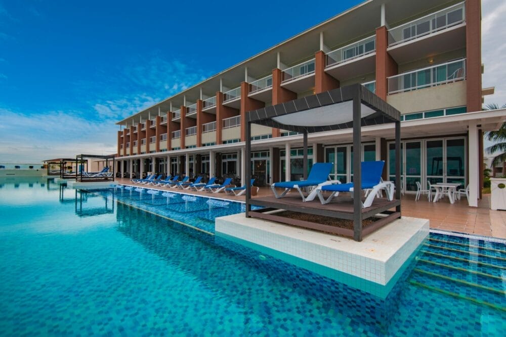 Il moderno Searesort Playa Vista Azul dispone di balconi che si affacciano su una grande piscina trasparente. Sedie a sdraio blu e una cabana ombreggiata invitano a rilassarsi a bordo piscina sotto il cielo azzurro.