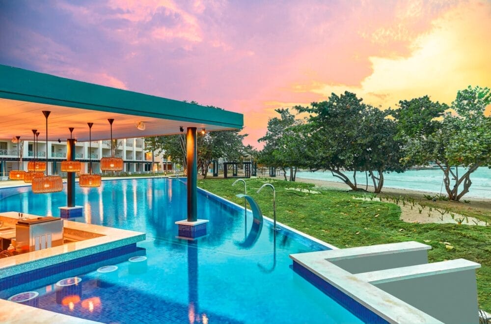 La lussuosa piscina con bar a bordo piscina del Royalton Negril Resort si affaccia su una spiaggia alberata al tramonto, dove le morbide sfumature rosa e arancioni illuminano il cielo e si riflettono sull'acqua.