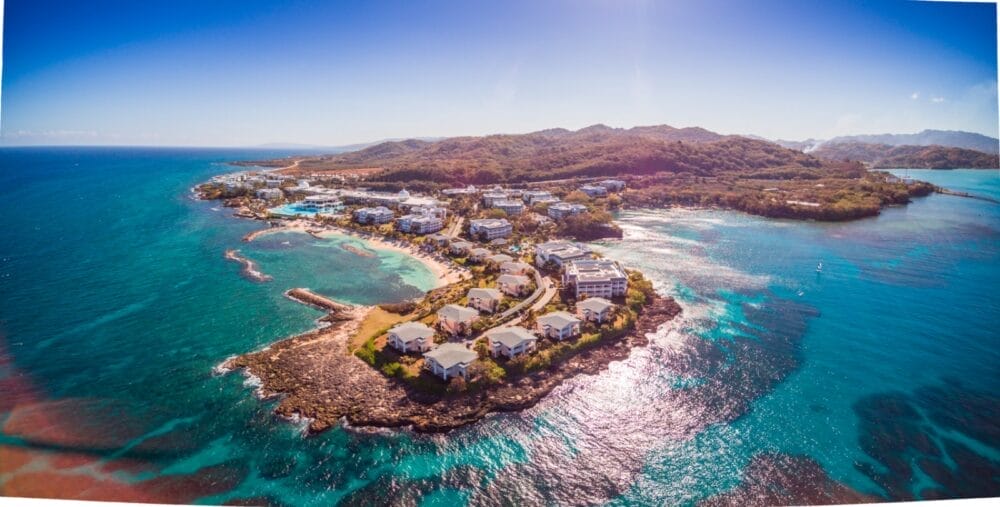 Veduta aerea del Grand Palladium Jamaica, una località costiera tropicale con edifici immersi in una vegetazione lussureggiante, acque turchesi e una costa rocciosa sotto un cielo azzurro e limpido.