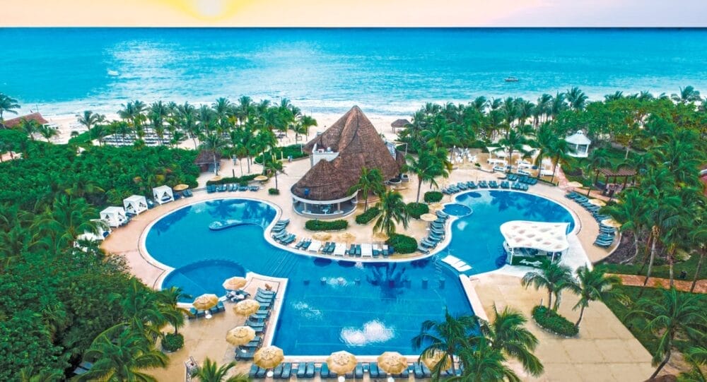 Vista aerea del Catalonia Playa Maroma, un resort sulla spiaggia tropicale con una grande piscina sinuosa, circondata da palme e ombrelloni, che si affaccia sulla spiaggia sabbiosa di Playa Maroma e sull'oceano turchese al tramonto.