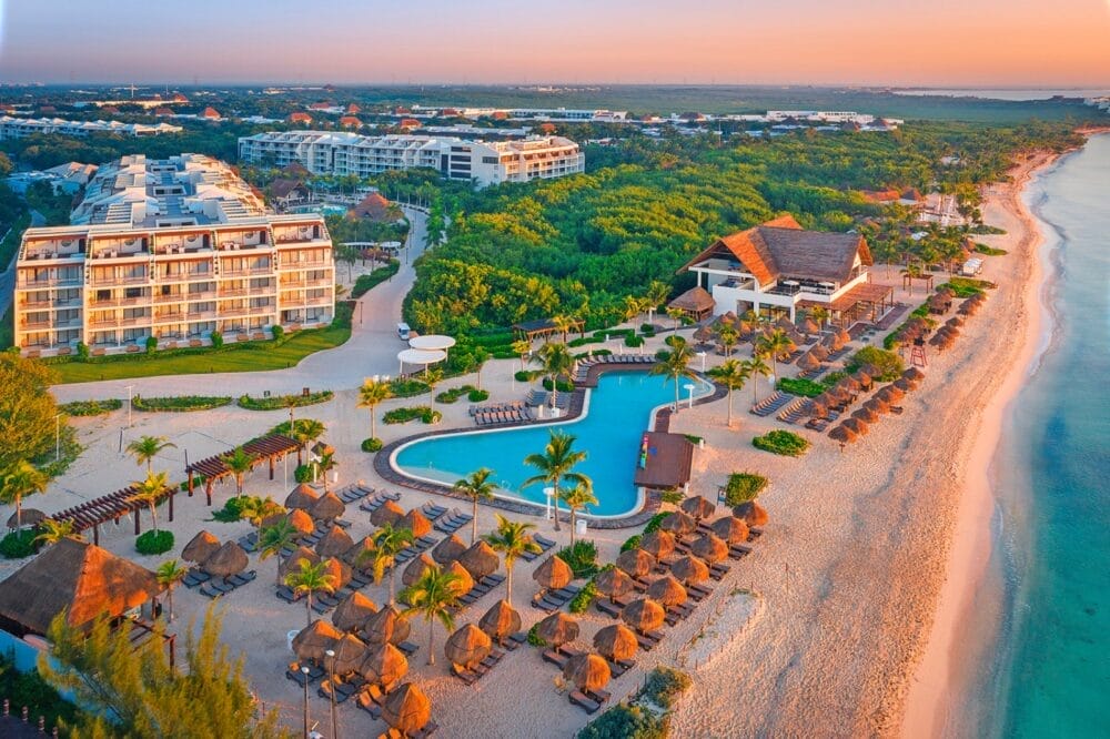 Veduta aerea dell'Ocean Riviera Paradise, uno splendido resort marittimo al tramonto, caratterizzato da una piscina centrale, palme, sedie a sdraio all'aperto con ombrelloni palapa, una spiaggia sabbiosa e grandi edifici dell'hotel circondati da una vegetazione lussureggiante.