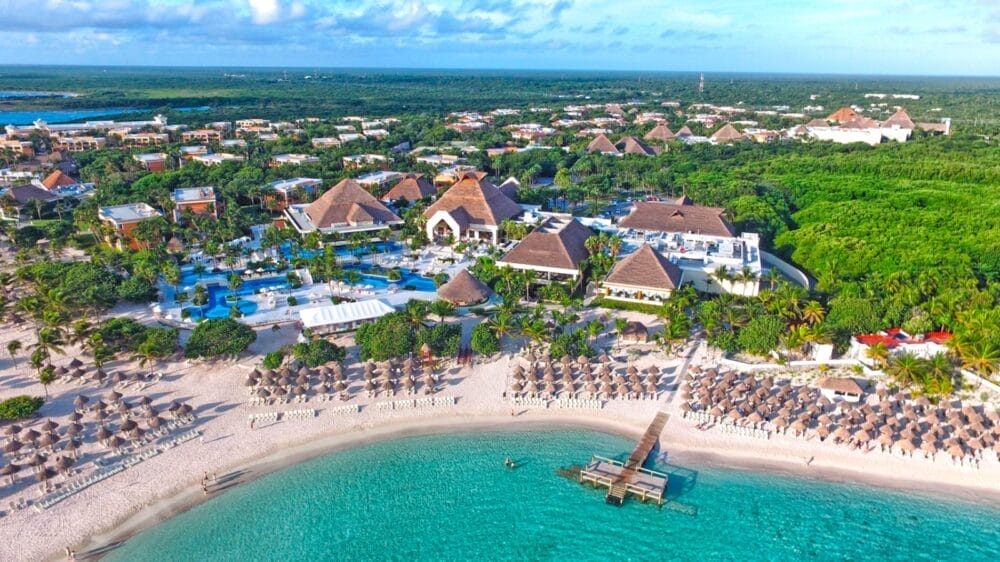 Vista aerea del resort sulla spiaggia tropicale Luxury Akumal, con edifici dal tetto di paglia, una piscina all'aperto, palme, file di sedie a sdraio e ombrelloni, un molo di legno, acqua turchese e una vegetazione lussureggiante vicino a Bahia Principe.