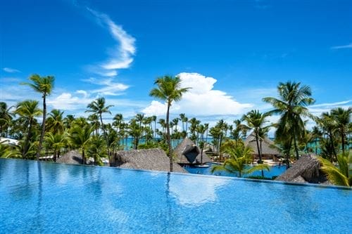 La piscina a sfioro blu chiaro del Barcelo Bavaro Palace si affaccia su palme, capanne con tetto di paglia e sull'oceano sotto un cielo luminoso e soleggiato con nuvole sparse: la vera atmosfera da resort all inclusive di Punta Cana.