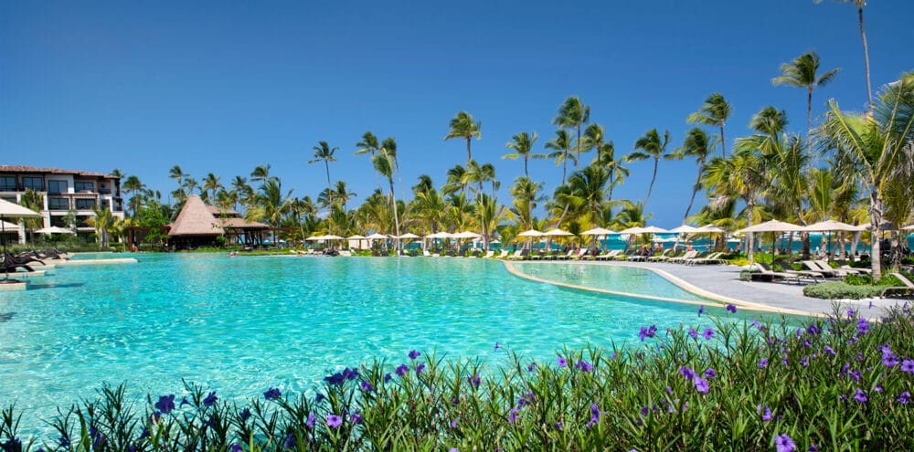 Una grande piscina trasparente circondata da palme, lettini e ombrelloni gialli sotto un cielo blu brillante al Lopesan Costa Bavaro, con fiori tropicali in primo piano ed edifici del resort sullo sfondo.