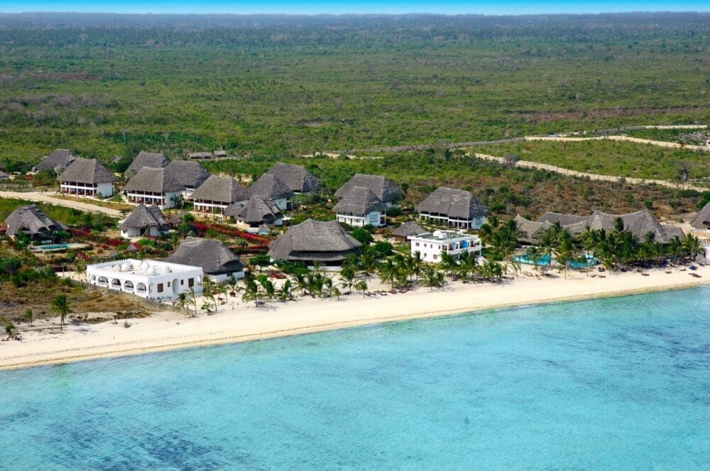 Vista aerea del resort Bravo Watamu sulla spiaggia, con edifici dai tetti di paglia e strutture bianche immerse nel verde, di fronte a un'acqua turchese e a una spiaggia sabbiosa. Sullo sfondo si estende una fitta foresta.