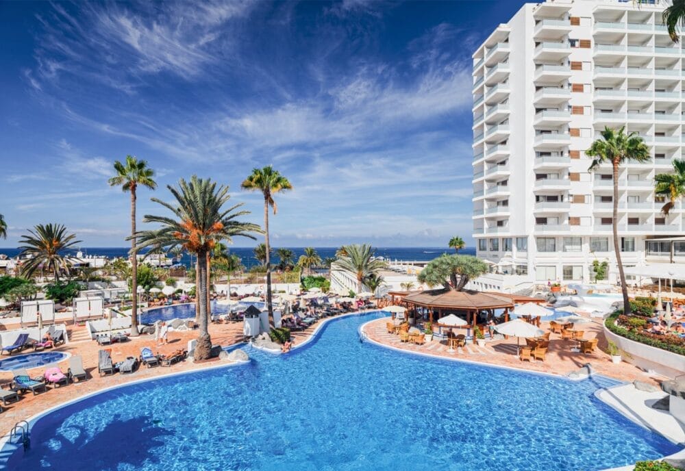 La grande piscina all'aperto del resort Seaclub H10 Gran Tinerfe è circondata da palme, lettini e ombrelloni, con gli ospiti che si rilassano e l'oceano e l'alto hotel bianco di Tenerife visibili sotto un cielo azzurro e limpido.