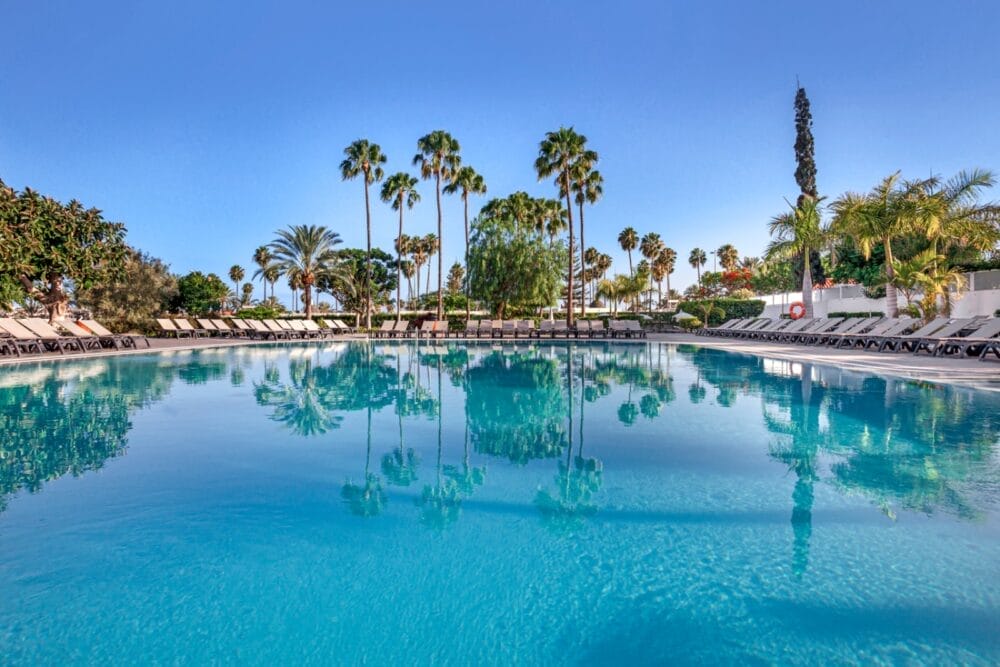 La piscina azzurra del Seaclub Barcelò Margaritas è circondata da palme e sedie a sdraio sotto un cielo luminoso e soleggiato, con gli alberi che si riflettono nell'acqua calma.