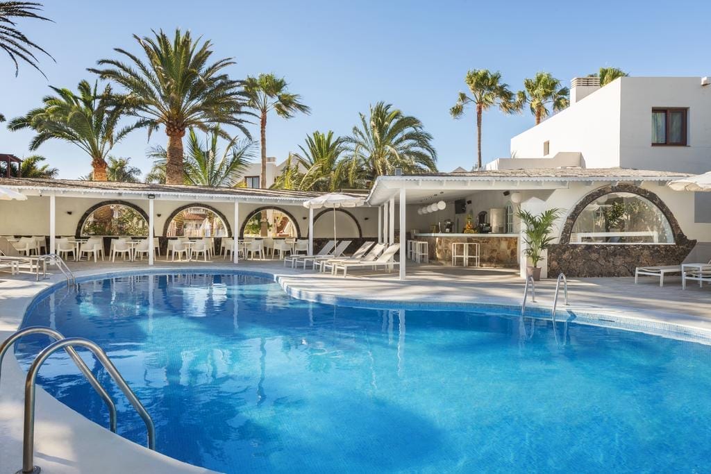 Godetevi la piscina all'aperto dell'Alua Suites Fuerteventura, un resort sul mare con sedie a sdraio, palme e un edificio bianco con archi. Rilassatevi al bar a bordo piscina sotto il patio ombreggiato in una giornata di sole a Fuerteventura.