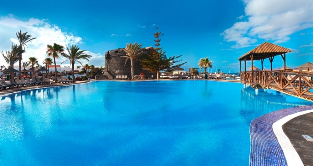Una grande e invitante piscina all'aperto sotto un cielo blu brillante al Barcelo Fuerteventura Seaclub, circondata da palme, sedie a sdraio e un gazebo in legno, con la storica torre di pietra del Castillo sullo sfondo.