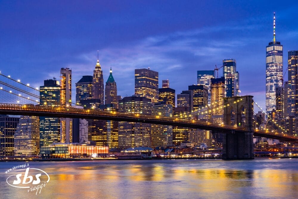 Il ponte di Brooklyn attraversa l'East River all'imbrunire, con lo skyline di Manhattan illuminato e il One World Trade Center sullo sfondo, creando uno splendido riflesso sull'acqua: una scena degna di un capolavoro di Bozza automatica.