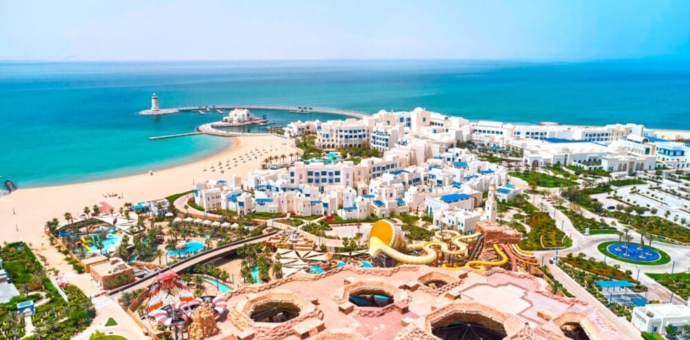 Vista aerea dell'Hilton Salwa Beach Resort con edifici bianchi, accenti blu, spiagge sabbiose, un porticciolo, scivoli d'acqua, palme e mare turchese sotto un cielo limpido, il tutto all'insegna del vero Seaclub Style.