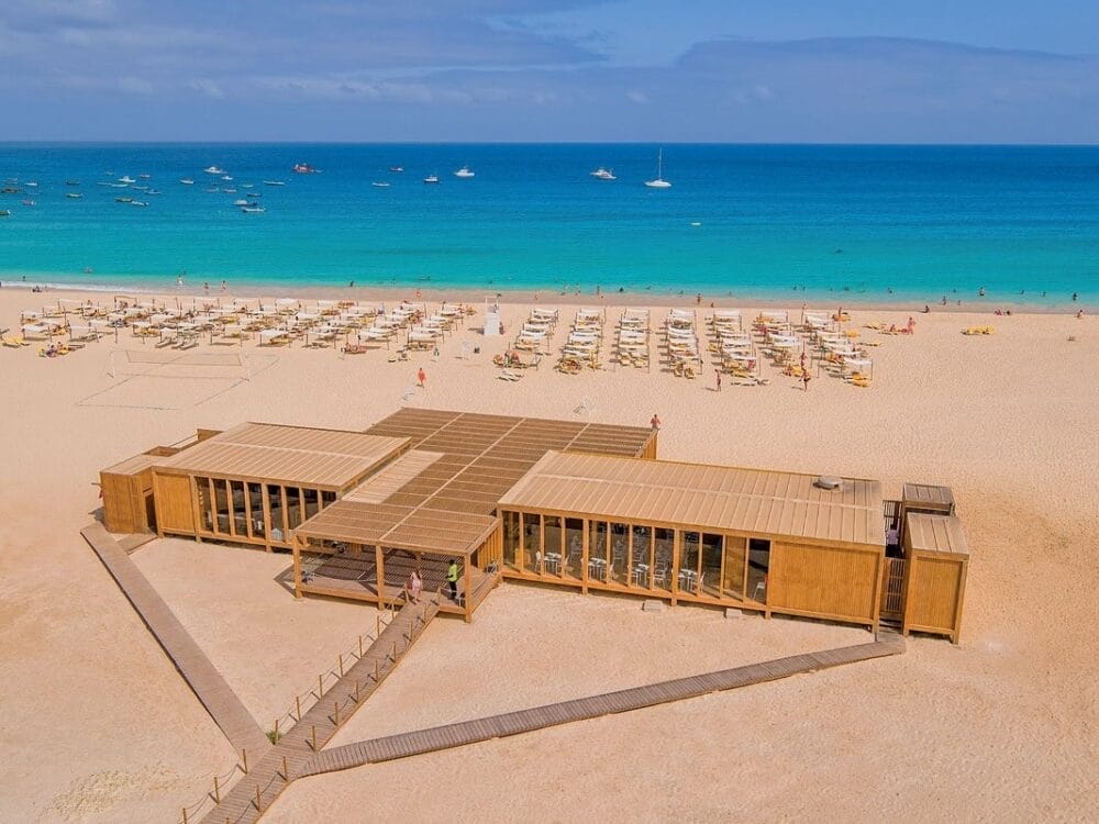 Vista aerea del Veraclub Oasis Salinas Sea, un moderno beach bar in legno su sabbia dorata, con file di lettini e ombrelloni affacciati su un mare turchese. Alcune persone si rilassano sulla spiaggia, mentre le barche vanno alla deriva nell'acqua sotto un cielo azzurro e limpido.