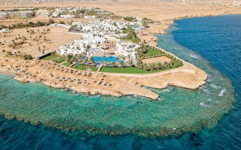 Vista aerea del resort Veraclub Sharm con edifici bianchi, prati verdi e palme su una costa rocciosa accanto a un'acqua limpida e turchese, delimitata dall'arido paesaggio desertico di Sharm el Sheikh.