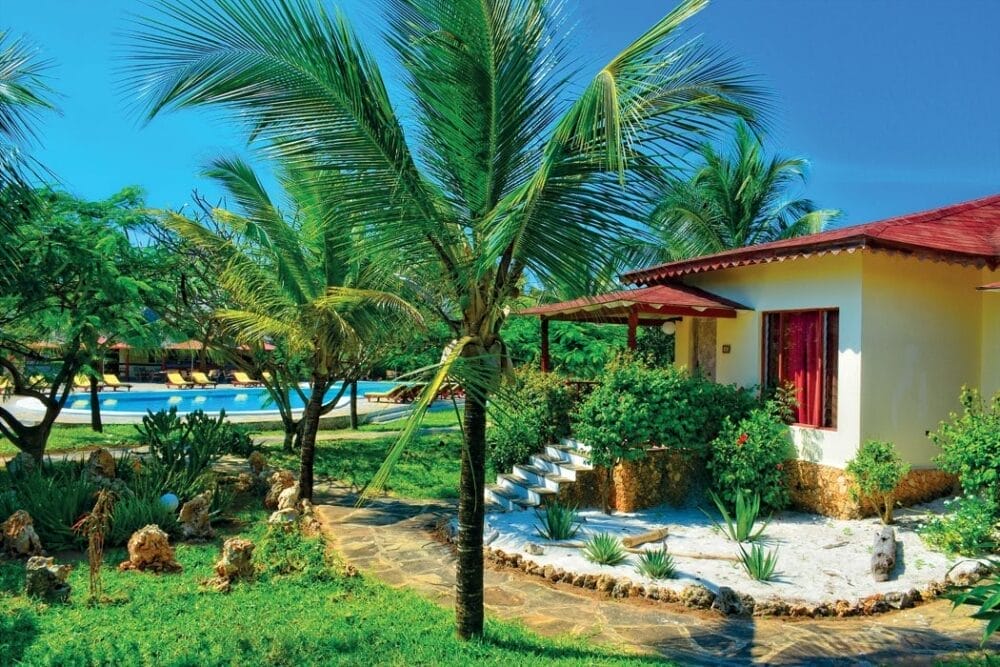 Un giardino tropicale con palme circonda un piccolo cottage giallo con tetto rosso e veranda ombreggiata al Veraclub Crystal Bay. Sullo sfondo, una piscina blu brilla tra la vegetazione lussureggiante sotto un cielo azzurro e limpido.