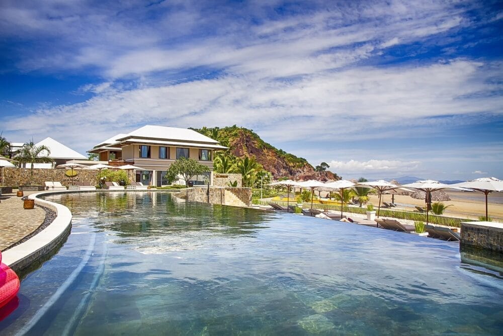 Resort di lusso Veraclub con piscina a sfioro che si affaccia sulla spiaggia sabbiosa di Palm Beach, lettini e ombrelloni, su un edificio moderno e una collina rocciosa, sotto un cielo azzurro con nuvole sparse.